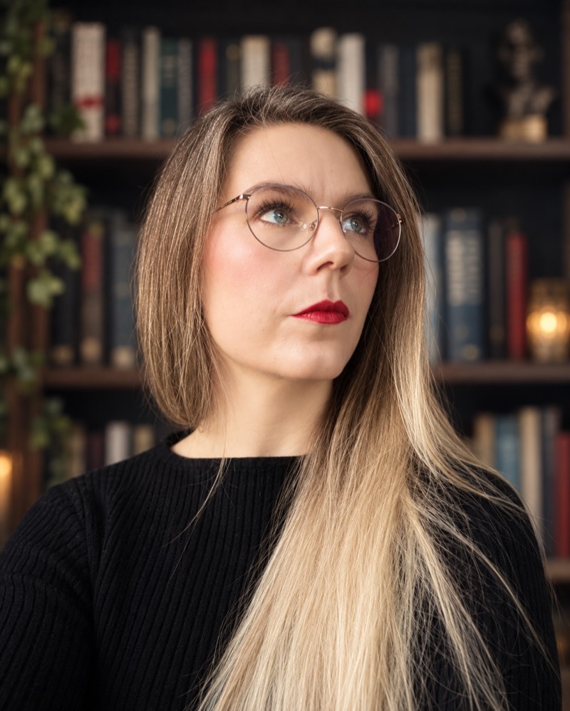 Porträt von Sarah Reichert: Sie steht vor einer dunklen Bücherwand, trägt eine feine Brille und einen schwarzen Rippenpullover und blickt an der Kamera vorbei nach oben.